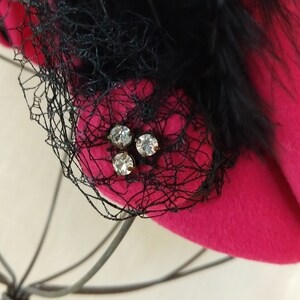 May include: A vibrant fuchsia hat with a black net veil and feather accents. The hat features a cluster of three sparkling rhinestones. The hat is a stylish accessory.