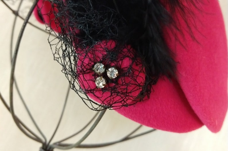 May include: A vibrant fuchsia hat with a black net veil and feather accents. The hat features a cluster of three sparkling rhinestones. The hat is a stylish accessory.