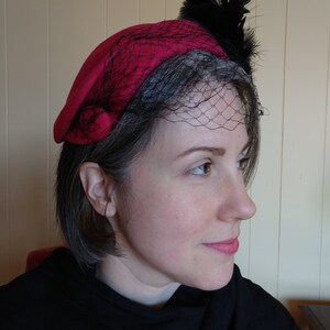 May include: A red beret-style hat with a black net veil and black feather accents. The hat is a vibrant shade of red and features a delicate net veil that drapes over the forehead. Black feathers are attached to the top of the hat.