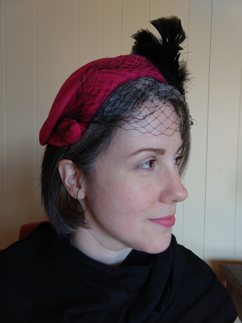 May include: A red beret-style hat with a black net veil and black feather accents. The hat is a vibrant shade of red and features a delicate net veil that drapes over the forehead. Black feathers are attached to the top of the hat.