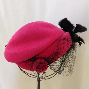 May include: A fuchsia-colored beret with a black veil and feather accent. The hat is displayed on a metal stand against a neutral background. The beret's design includes a draped crown and a delicate net veil.