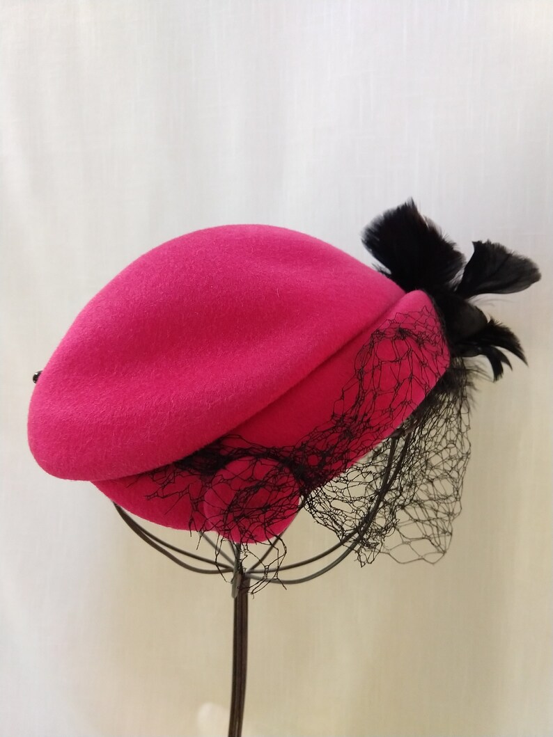 May include: A fuchsia-colored beret with a black veil and feather accent. The hat is displayed on a metal stand against a neutral background. The beret's design includes a draped crown and a delicate net veil.