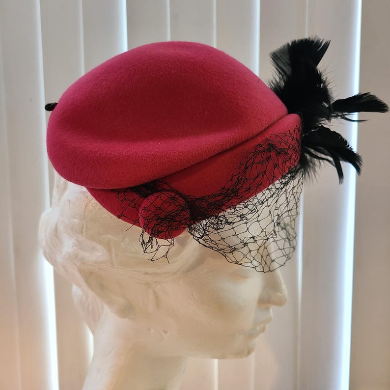 May include: A bright red hat with a rounded crown, adorned with black feathers and a black net veil. The hat has a small, decorative button on the side. This vintage-style hat is suitable for special occasions.