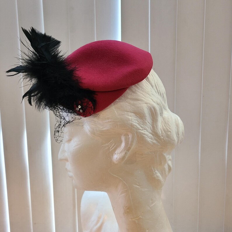 May include: A burgundy beret-style hat with black feather accents and a small decorative embellishment. The hat features a delicate black netting and is displayed on a mannequin head. The hat is a stylish accessory.