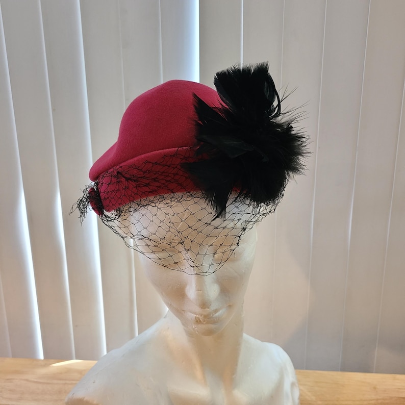 May include: A red cloche hat with a black feather accent and a black netting veil. The hat is made of a felt-like material and has a rounded crown and a small brim. The veil drapes over the front of the hat.