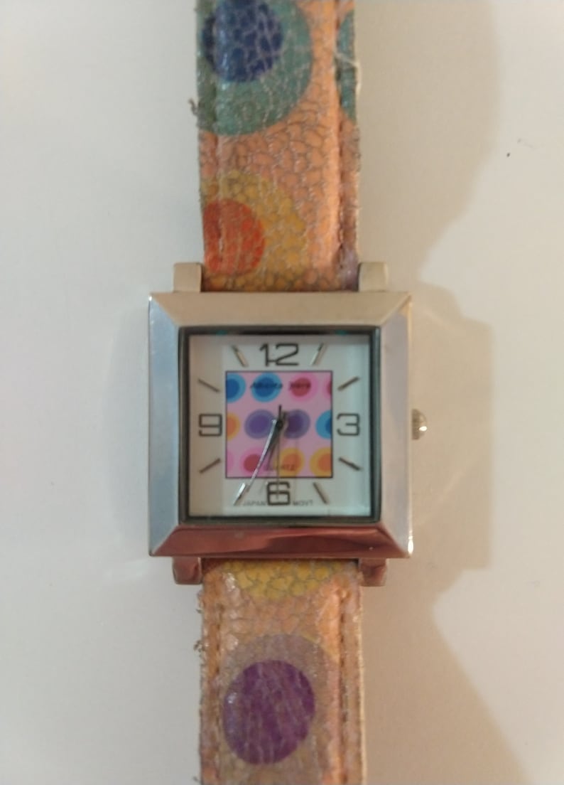 May include: A silver square watch with a white face and colorful polka dot design. The watch has a brown leather band with a colorful polka dot pattern.