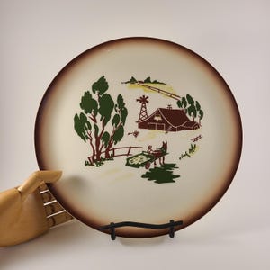 May include: A round ceramic plate with a cream-colored center and a wide, dark brown rim. The center features a graphic illustration of a farm scene with a brown barn, a windmill, green trees, and a horse pulling a cart filled with hay.