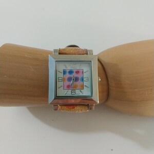 May include: A silver square watch with a white face and colorful polka dot design. The watch has a brown leather band with a floral pattern.