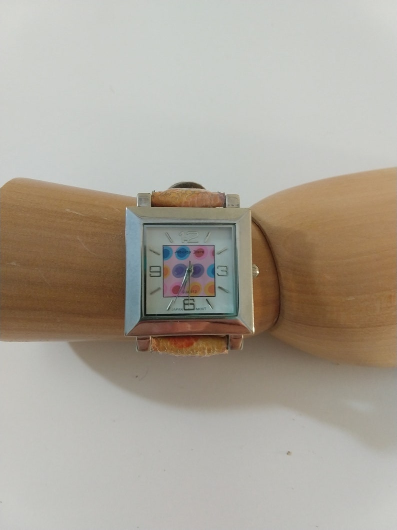May include: A silver square watch with a white face and colorful polka dot design. The watch has a brown leather band with a floral pattern.