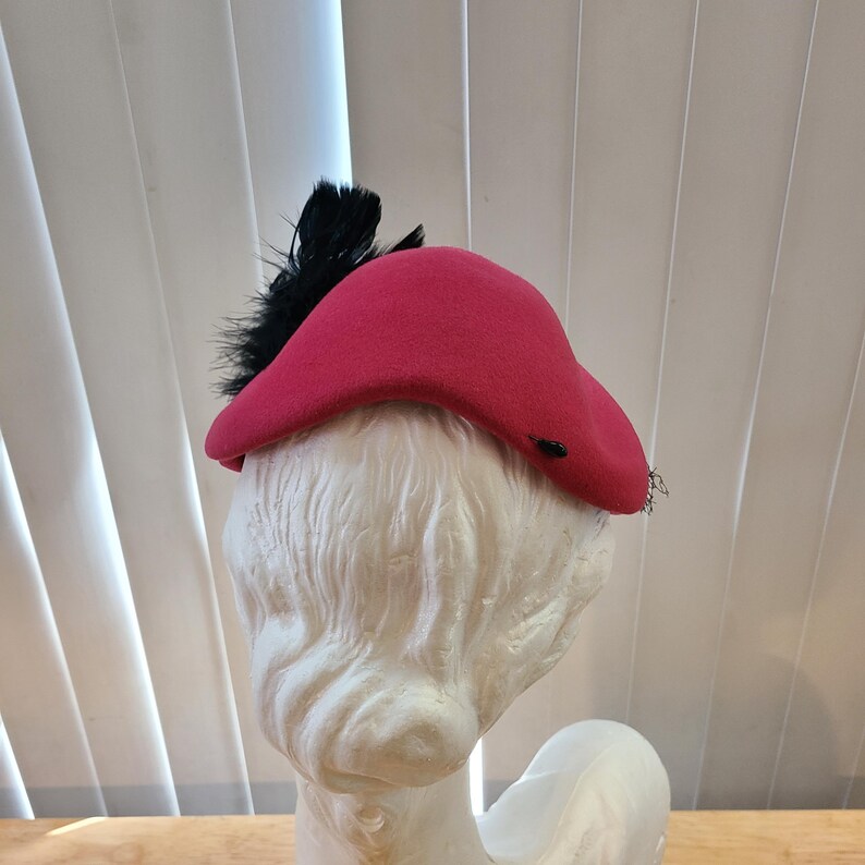 May include: A bright red hat with a curved brim and a cluster of black feathers. The hat is displayed on a white mannequin head, showcasing its shape and design. The hat's vibrant color and feather accent create a striking visual.
