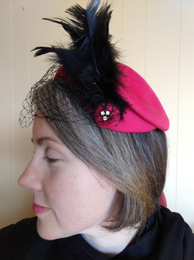 May include: A fuchsia-colored hat with black feathers and a black netting veil. The hat is adorned with a cluster of small, sparkling rhinestones. The hat is a stylish accessory.