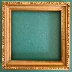 May include: A square, antique gold picture frame with ornate detailing. The frame has a rectangular opening and is set against a teal green background. The frame's edges feature a repeating scroll design.