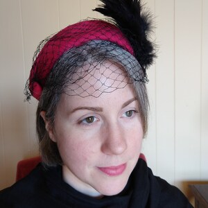 May include: A vintage-style hat with a deep red crown and a gray brim. The hat features a black netting veil and is adorned with a cluster of black feathers. The hat is a classic accessory.