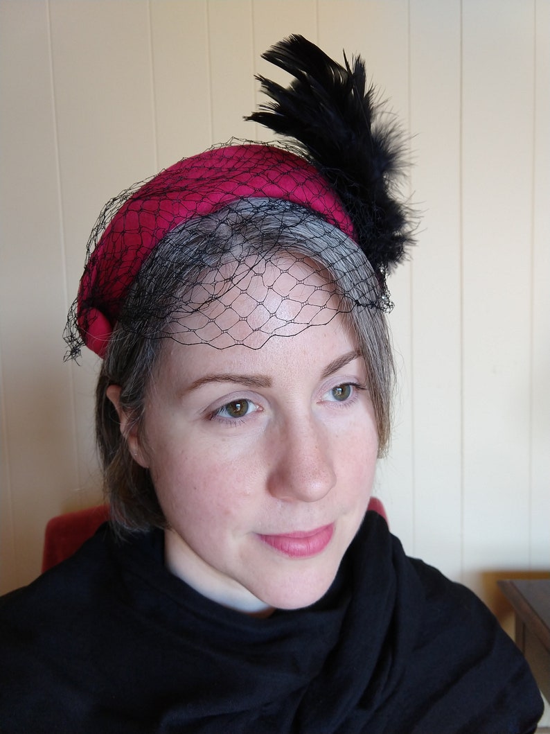 May include: A vintage-style hat with a deep red crown and a gray brim. The hat features a black netting veil and is adorned with a cluster of black feathers. The hat is a classic accessory.