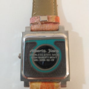 May include: A silver watch with a square face and a brown leather band. The watch face has a turquoise border and the text "Alberta Flora" and "Stainless Steel Back High Quality Movt" is printed on the face. The band has a pink and orange pattern.