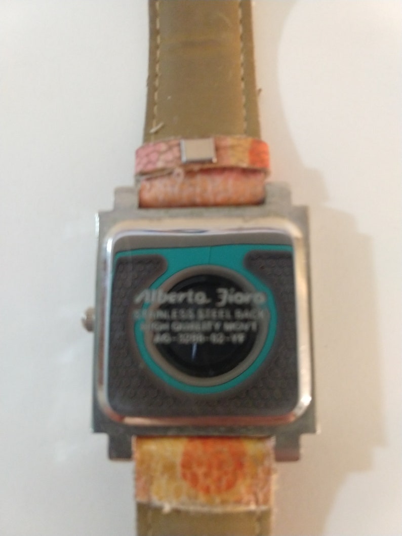 May include: A silver watch with a square face and a brown leather band. The watch face has a turquoise border and the text "Alberta Flora" and "Stainless Steel Back High Quality Movt" is printed on the face. The band has a pink and orange pattern.