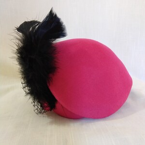 May include: A bright pink hat with a rounded crown and a black feather and netting accent. The hat is a vibrant color and appears to be made of felt or a similar material. The black feathers add a touch of elegance.