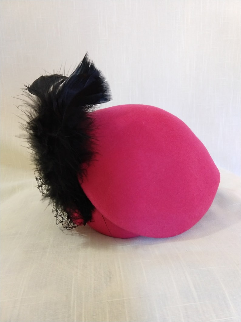 May include: A bright pink hat with a rounded crown and a black feather and netting accent. The hat is a vibrant color and appears to be made of felt or a similar material. The black feathers add a touch of elegance.