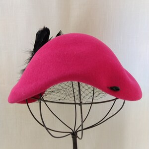 May include: A vibrant fuchsia hat with a curved brim and a cluster of black feathers. The hat features a small, dark button detail and is displayed on a wire hat stand. The hat's design is reminiscent of vintage fashion.