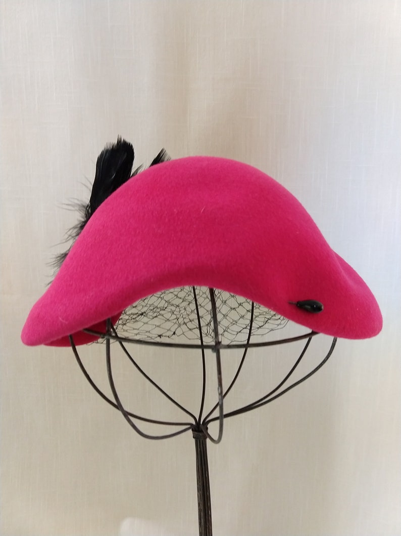 May include: A vibrant fuchsia hat with a curved brim and a cluster of black feathers. The hat features a small, dark button detail and is displayed on a wire hat stand. The hat's design is reminiscent of vintage fashion.