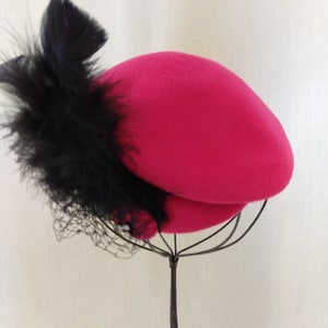 May include: A bright pink hat with a black feather and netting accent. The hat is displayed on a wire stand against a white background. The hat's design is a classic, elegant style.