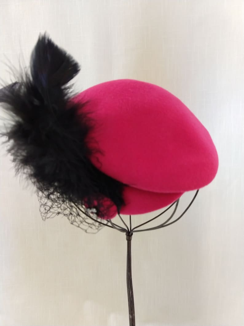 May include: A bright pink hat with a black feather and netting accent. The hat is displayed on a wire stand against a white background. The hat's design is a classic, elegant style.