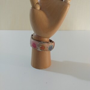 May include: A wooden hand model wearing a pink and multi-colored patterned watch band with a silver buckle.