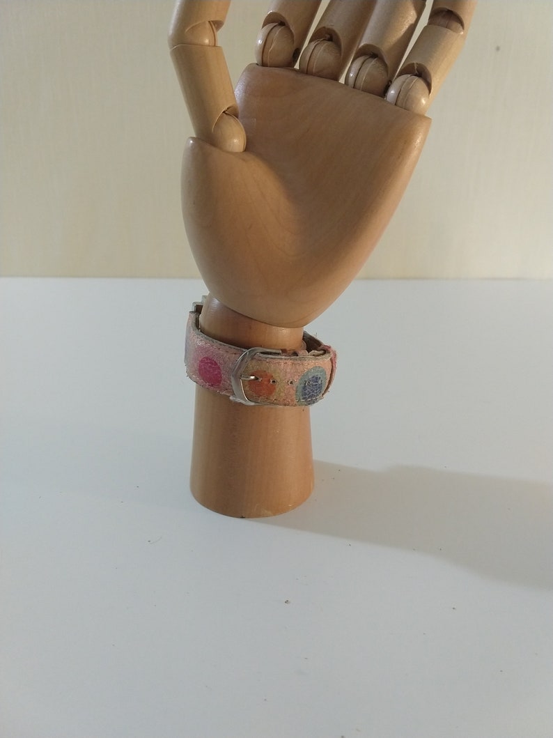 May include: A wooden hand model wearing a pink and multi-colored patterned watch band with a silver buckle.