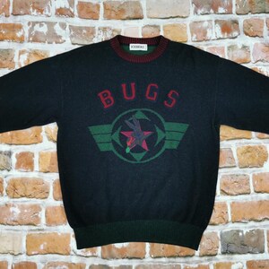 Könnte beinhalten: Schwarzer Pullover mit einer grünen, roten und weißen Grafik eines Flugzeugs und dem Wort "BUGS" in Rot.
