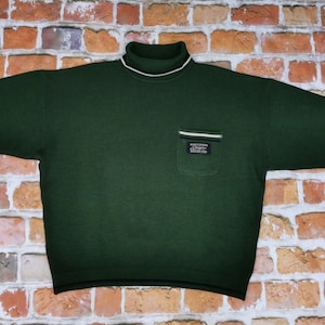 Puede incluir: Un suéter de cuello alto verde oscuro con un pequeño bolsillo en el lado izquierdo. El bolsillo tiene un ribete blanco y una etiqueta negra que dice "Just Cavalli - Restricted".