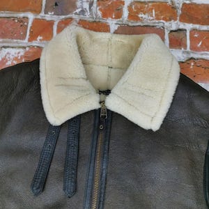 Pu&ograve; includere: Primo piano di una giacca in pelle marrone con colletto in montone color crema. La giacca ha una chiusura con cerniera e cinturini in pelle nera. Uno stile classico, ideale per il freddo.
