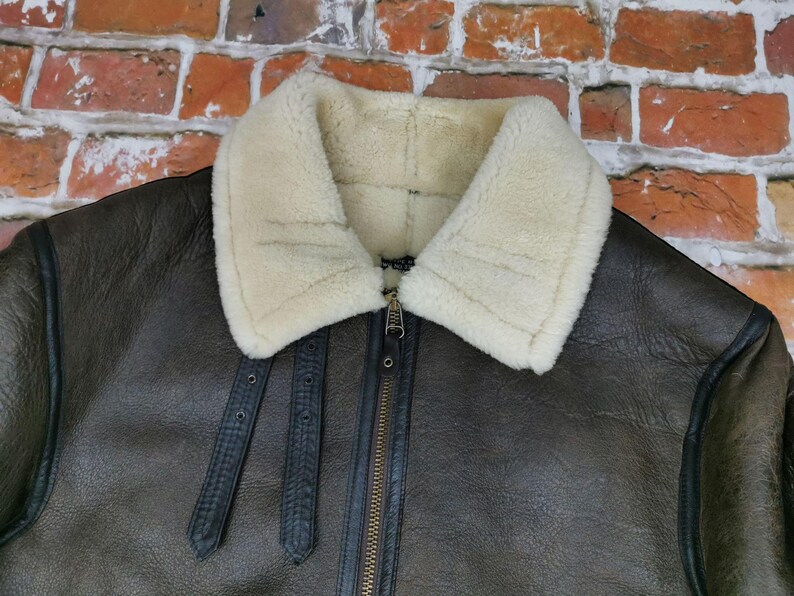 Pu&ograve; includere: Primo piano di una giacca in pelle marrone con colletto in montone color crema. La giacca ha una chiusura con cerniera e cinturini in pelle nera. Uno stile classico, ideale per il freddo.