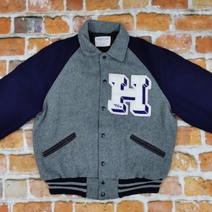 Könnte beinhalten: Eine graue und marineblaue Collegejacke mit einem großen weißen Buchstaben "H" auf der Vorderseite. Die Jacke hat einen Reißverschluss und einen gerippten Kragen, Bündchen und Saum.