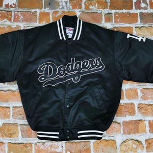 Puede incluir: Una chaqueta bomber de satén negro con el logotipo de Dodgers bordado en blanco en el pecho y un logotipo de LA en blanco en la manga izquierda. La chaqueta tiene cuello y puños acanalados con rayas blancas, cierre de botones a presión y dos bolsillos.