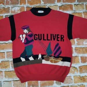 Può includere: Maglione rosso a maniche lunghe con bordino a righe bianche e nere. Il maglione presenta una grafica colorata di una persona che naviga su una barca con la scritta "GULLIVER" stampata sul davanti.