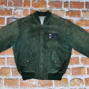 Peut inclure: Une veste bomber en daim vert foncé avec une fermeture éclair et un petit patch sur le côté gauche. La veste a un col, des poignets et une ceinture côtelés.