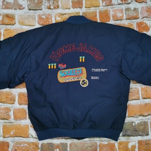 May include: A navy blue bomber jacket with the text "HOME JAMES" and "The SQUEEZE CHEVIGNON 5¢" embroidered on the back.