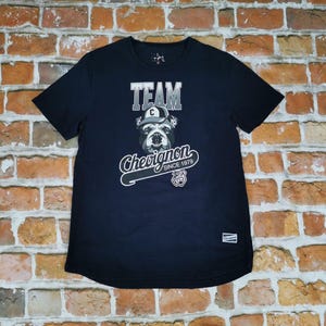 Puede incluir: Una camiseta azul marino con un gráfico que presenta la palabra "TEAM" sobre un bulldog con gorra. La camiseta también muestra el texto "Chevignon" y "SINCE 1979" en una fuente estilizada. Una pequeña etiqueta es visible en la parte inferior derecha.