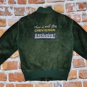 Può includere: Una giacca bomber in pelle scamosciata verde scuro con un logo ricamato giallo e bianco che recita "There is only one Chevignon Exclusive!"