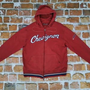 Puede incluir: Sudadera roja con cremallera y bolsillos delanteros. La palabra "Chevignon" está bordada en el pecho en escritura blanca. Los puños y el dobladillo presentan rayas azules y blancas. La sudadera con capucha se muestra contra una pared de ladrillos.
