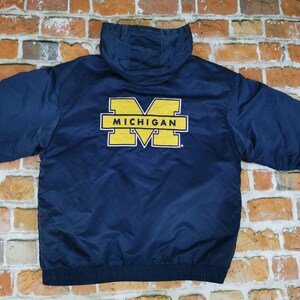 May include: A navy blue hooded jacket featuring a large yellow block letter M logo on the back with the word MICHIGAN printed in black across the center of the letter. The jacket has long sleeves, a hood, and a ribbed hem at the bottom edge.