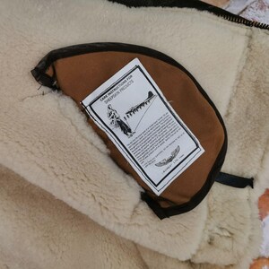 Pu&ograve; includere: Primo piano dell'interno di una giacca in pelle di pecora. La giacca presenta una fodera color crema e soffice e una tasca marrone con un'etichetta con le istruzioni per la cura. L'etichetta include un disegno di una persona e di un aereo.