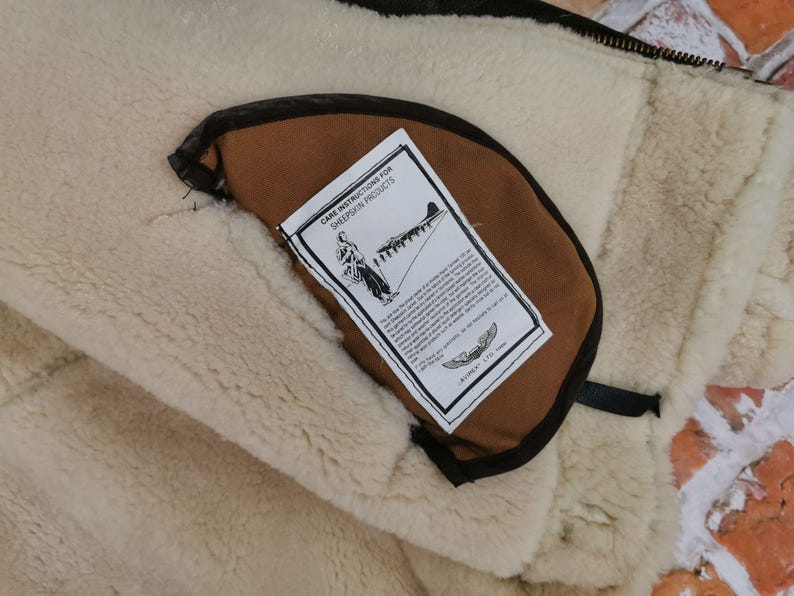 Pu&ograve; includere: Primo piano dell'interno di una giacca in pelle di pecora. La giacca presenta una fodera color crema e soffice e una tasca marrone con un'etichetta con le istruzioni per la cura. L'etichetta include un disegno di una persona e di un aereo.