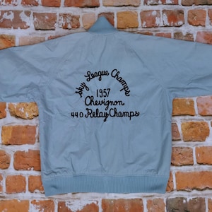 May include: A light blue jacket with a black embroidered patch that says "Sky League Champs 1957 Chevignon 440 Relay Champs".
