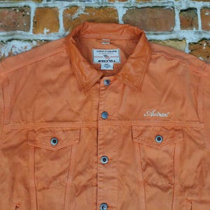 Puede incluir: Chaqueta vaquera naranja con cuello, cierre de botones y dos bolsillos en el pecho. La palabra "Autrex" est&aacute; bordada en blanco. La chaqueta est&aacute; hecha de un material resistente.
