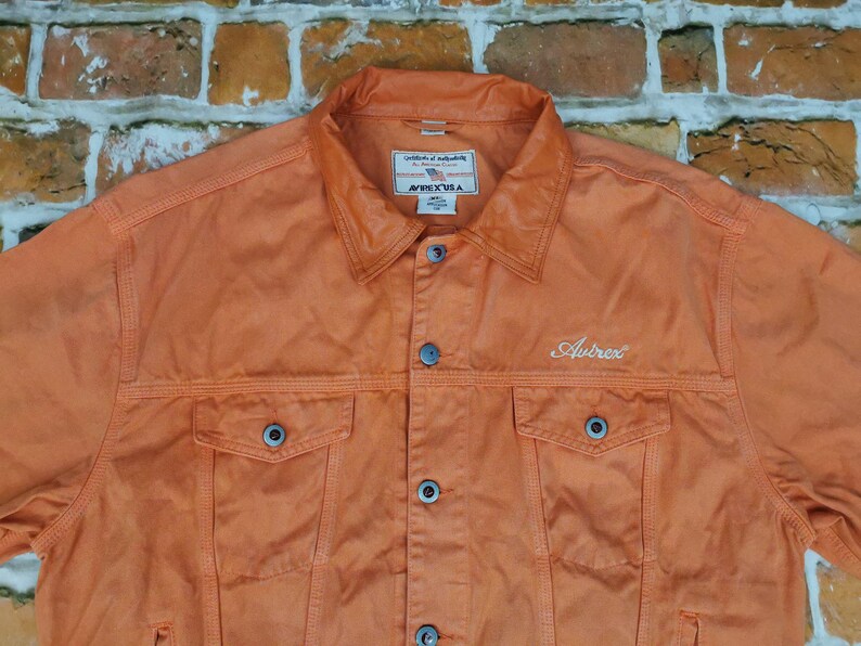 Puede incluir: Chaqueta vaquera naranja con cuello, cierre de botones y dos bolsillos en el pecho. La palabra "Autrex" est&aacute; bordada en blanco. La chaqueta est&aacute; hecha de un material resistente.