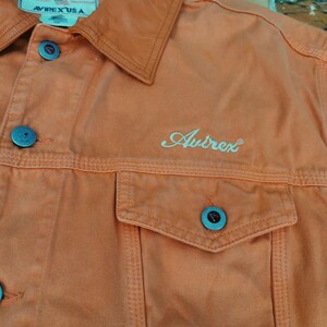 Puede incluir: Primer plano de una chaqueta vaquera naranja con cuello y dos bolsillos en el pecho. La chaqueta tiene botones turquesa y la palabra "Avirex" bordada en la parte delantera. La etiqueta dice "Avirex USA".