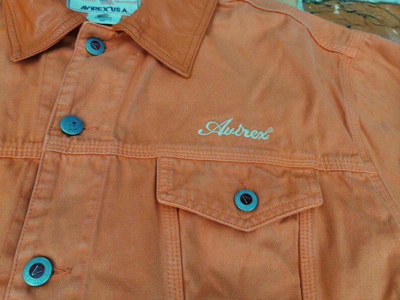 Puede incluir: Primer plano de una chaqueta vaquera naranja con cuello y dos bolsillos en el pecho. La chaqueta tiene botones turquesa y la palabra "Avirex" bordada en la parte delantera. La etiqueta dice "Avirex USA".