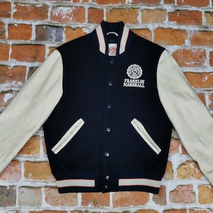 Op de afbeelding: Een donkerblauw en crème varsity jack met een wit geborduurd logo met de tekst "FRANKLIN MARSHALL".