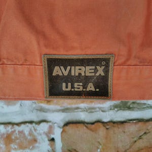 Puede incluir: Primer plano de una chaqueta naranja con el parche del logotipo "AVIREX U.S.A.". La chaqueta tiene dos botones plateados y una costura. El fondo es una pared de ladrillos.